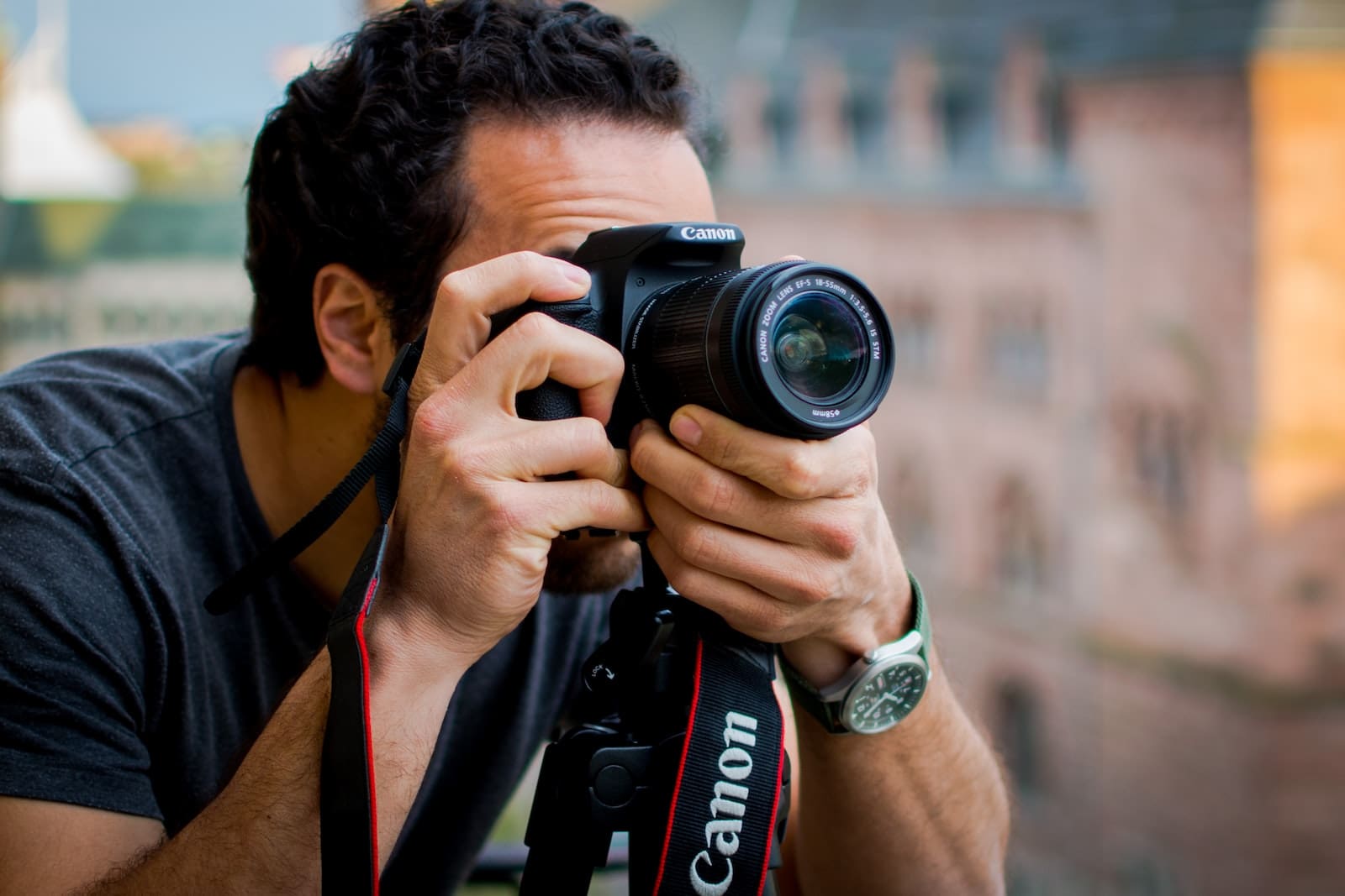 An enthusiastic photographer is focused on capturing the moment with a Canon DSLR camera, the strap clearly visible. The individual is wearing a dark shirt and a wristwatch, suggesting a casual yet prepared approach to photography. The blurred background hints at an urban environment, providing context for an outdoor photo shoot.