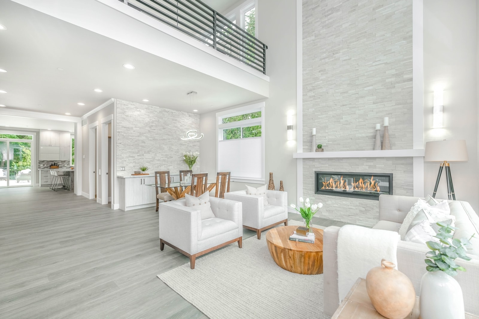 A spacious, modern living room bathed in natural light, featuring high ceilings and a contemporary design. The room is elegantly furnished with white armchairs, a round wooden coffee table, and a stone fireplace, creating a serene and welcoming atmosphere. In the background, an open-concept kitchen and dining area are visible, enhancing the room's airy feel.