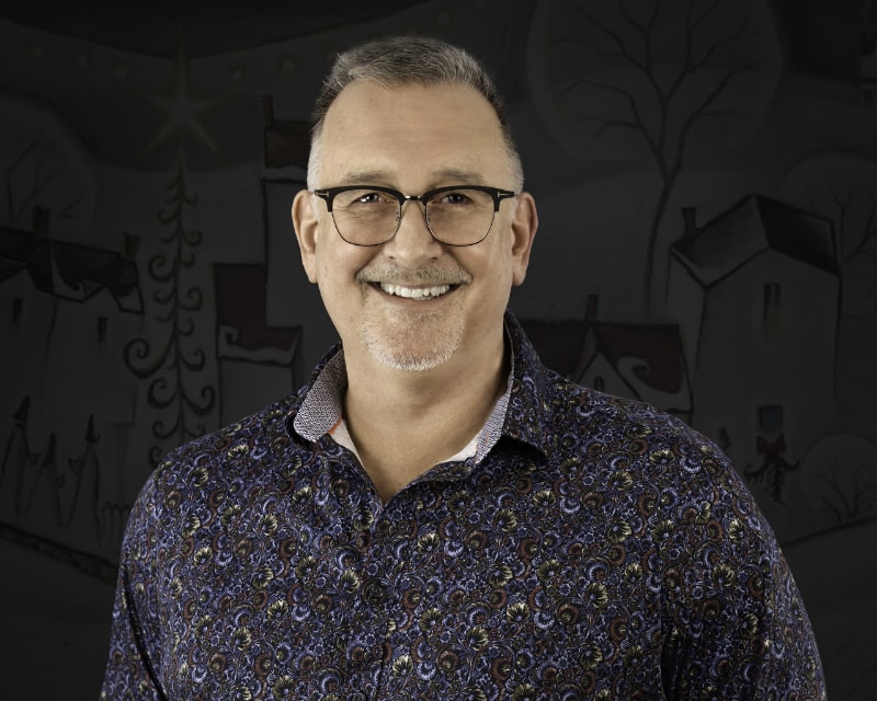 Dale Bryant, smiling in a paisley shirt and glasses, in front of a whimsical village backdrop.