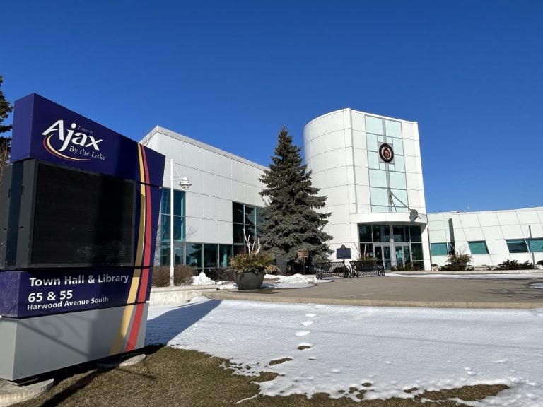 Ajax Real Estate: The image captures the exterior of the Ajax Town Hall and Library on a bright sunny day with clear blue skies. A large sign featuring the town's name, "Ajax - By the Lake," and address "65 & 55 Harwood Avenue South" is prominently displayed in the foreground. There's a dusting of snow on the lawn, and a mature evergreen tree stands near the modern glass-fronted building.