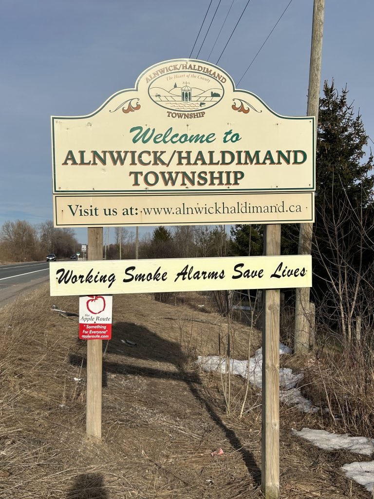Alnwick/Haldimand Sign, displaying welcoming lettering on a wooden board with a rustic charm, set against a backdrop of dense foliage in Ontario.