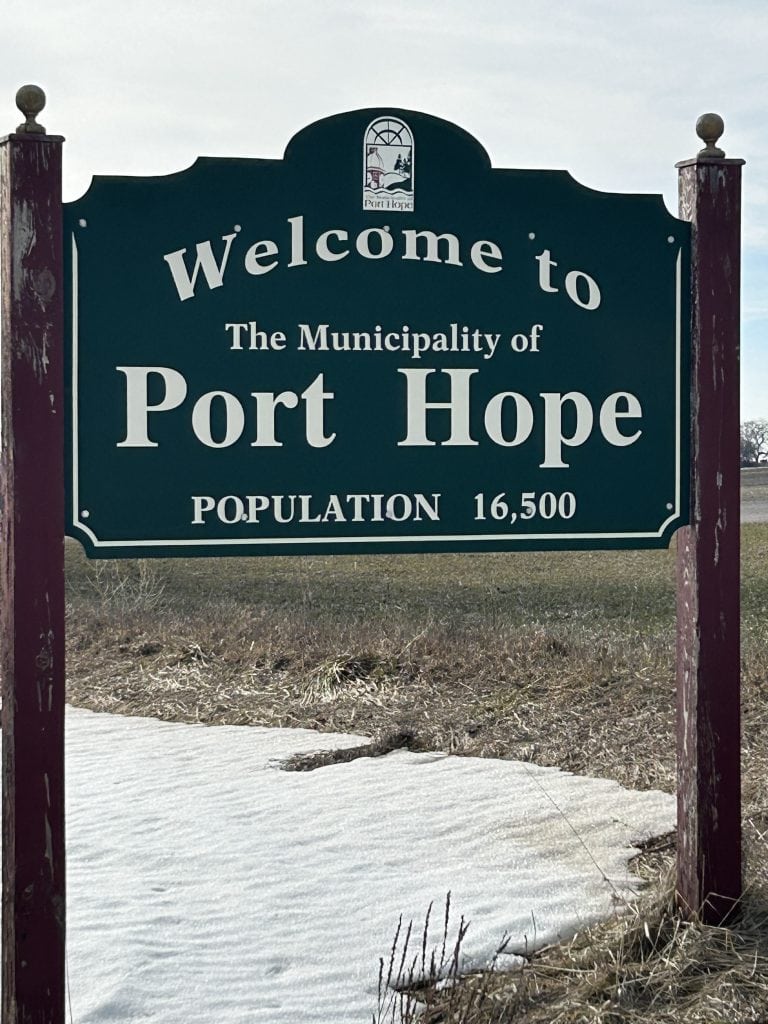 The Port Hope Sign features artistic lettering on a heritage-style wooden frame, nestled among lush greenery, inviting visitors into this vibrant, historic community in Ontario.