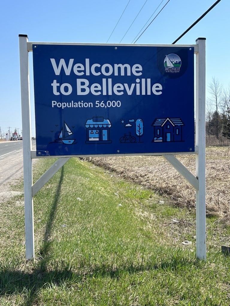 The Belleville Sign, prominently featuring bold, welcoming lettering atop a brick base, with a backdrop of lush trees and a clear blue sky, capturing the essence of this vibrant Ontario city.