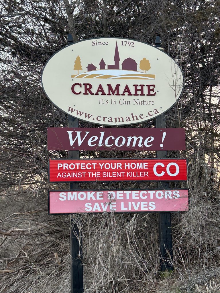 The Cramahe Sign is elegantly presented with a rustic wooden backdrop and bold lettering, flanked by Canadian flags and natural scenery, welcoming visitors to this historic community in Ontario.
