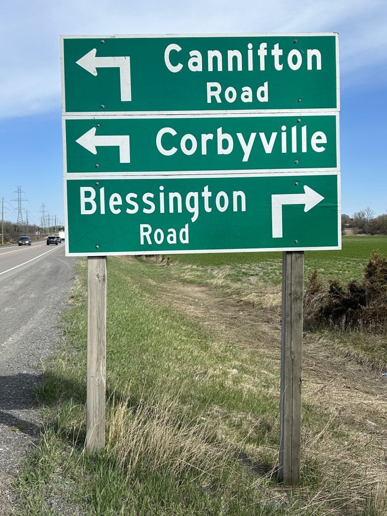 The Thurlow Road sign, displayed prominently against a rural backdrop, featuring bold lettering on a classic wooden frame, surrounded by lush foliage and clear skies, marking a scenic route in Ontario.
