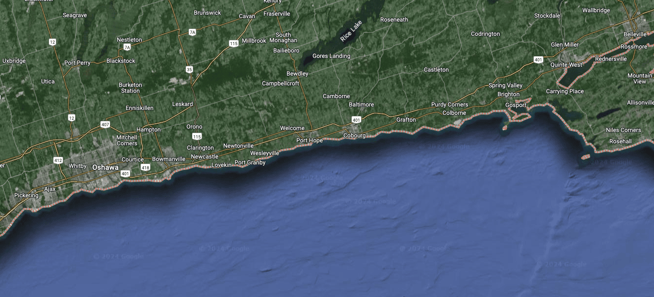 A satellite map view of a coastal region, with the names of various places visible such as Pickering, Ajax, Whitby, Oshawa, Clarington, Port Hope, Cobourg, and further east towards Belleville. Major highways like the 401 and 407 are marked, alongside smaller roads and towns set in a backdrop of green indicating vegetation and blue for the adjacent lake or sea.